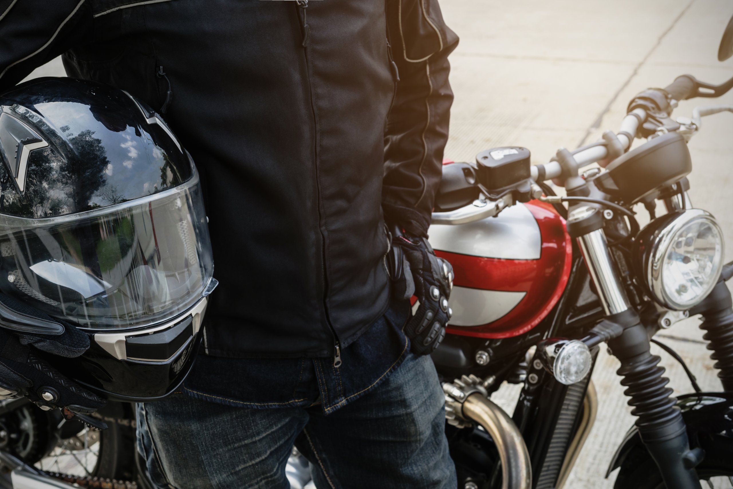 Close up of a person holding a helmet and wearing a leather jacket stands next to a motorcycle.