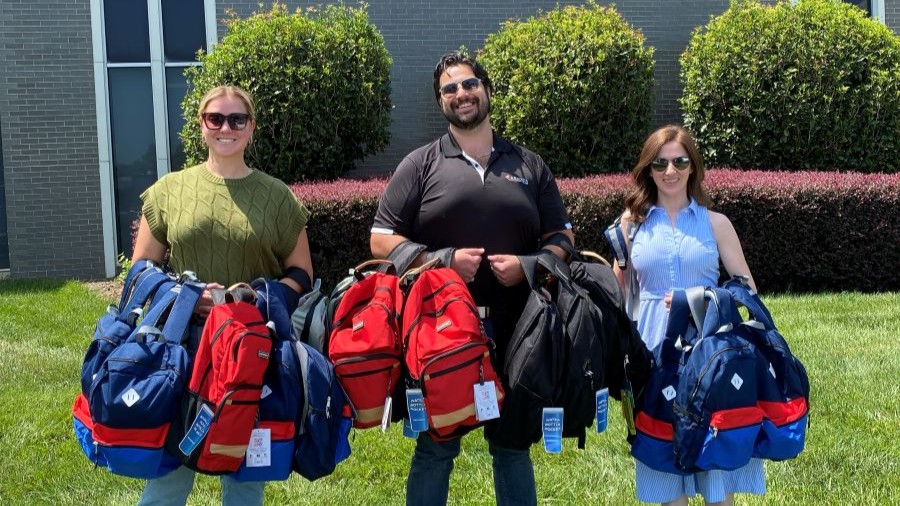 Anslee, Tim and Isabella stand outdoors holding various backpacks.